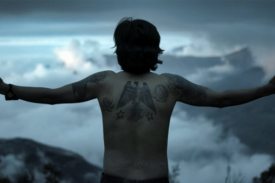 still from film; a boy with a back tattoo stands with arms outspread over a mountainous landscape